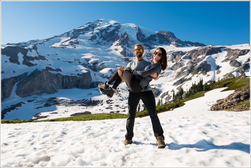 man holding fiancee off the snow with both arms and mt rainier in background