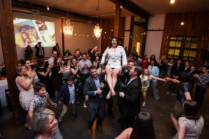 Woman sitting on chair lifted high during horah at her son's bat mitzvah