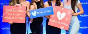 Four women from Make-A-Wish hold up signs in front of a step and repeat at the Westin in Seattle.