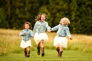 Three girls with jean jackets, white dresses, and brown cowboy boots running while holding hands and smiling