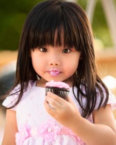 Asian girl holding chocolate cupcake with pink frosting in hand with pink frosting on lips while looking at camera