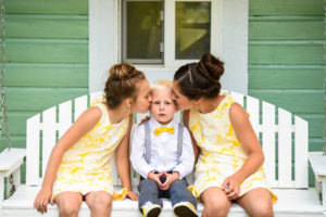 Two sisters wearing yellow dresses kissing brother with bowtie in between them sitting on a white bench.
