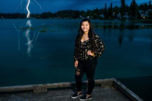 Girl posing for her senior photos in front of a lake with lightning in the background