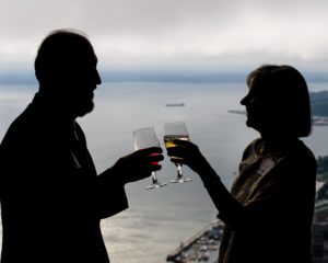 A man and woman cling glasses together in front of Columbia Tower windows overlooking Puget Sound