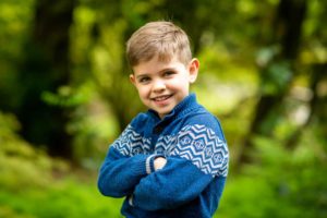 Boy in blue sweater with crossed arms smiling in forest