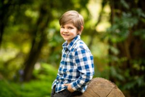 Boy with hands in pocket leaning against log smiling in forest