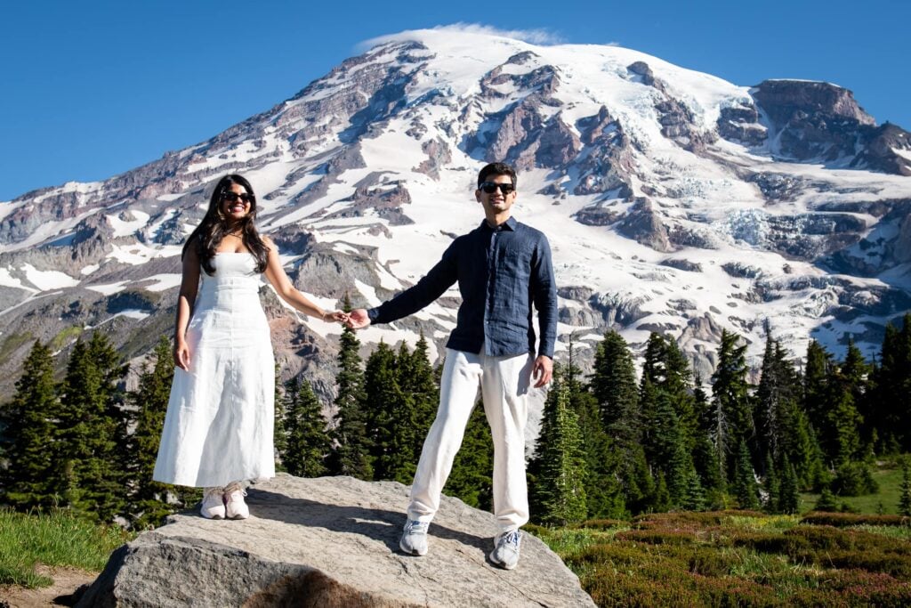 Nicely dressed couple standing on rock wearing sunglasses and holding hands with mt. rainier in the background