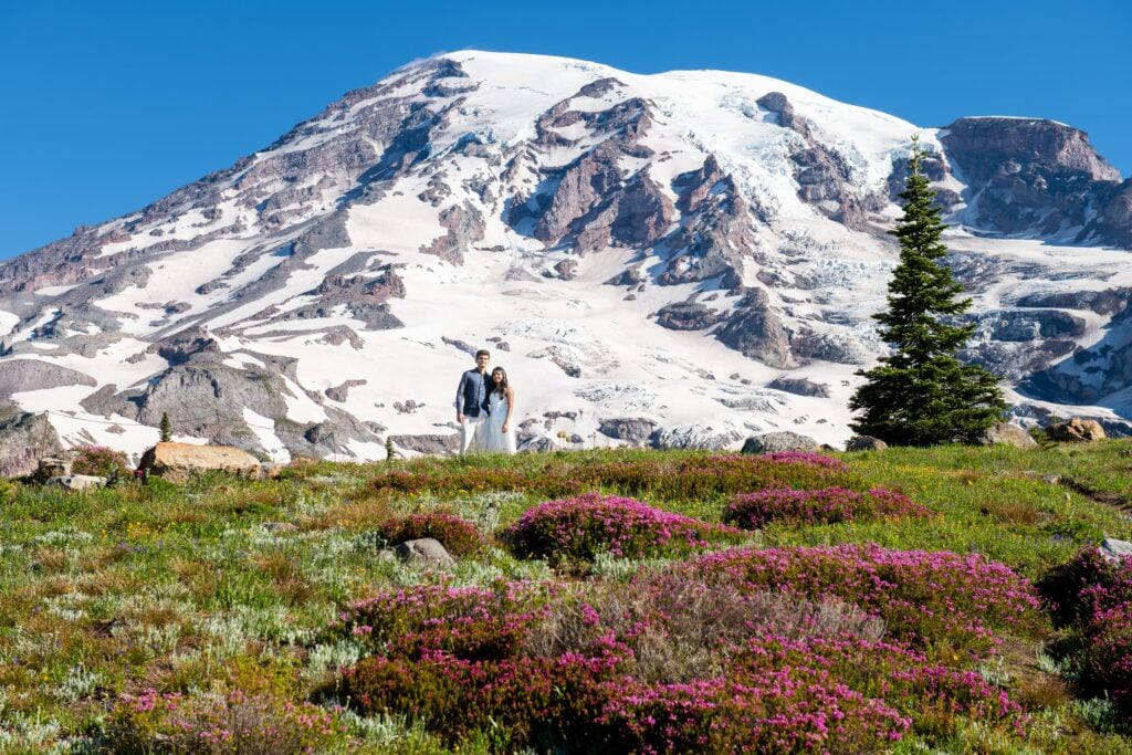 Engaged couple with arms around each other facing camera with wildflowers around them and mt. rainier in the background