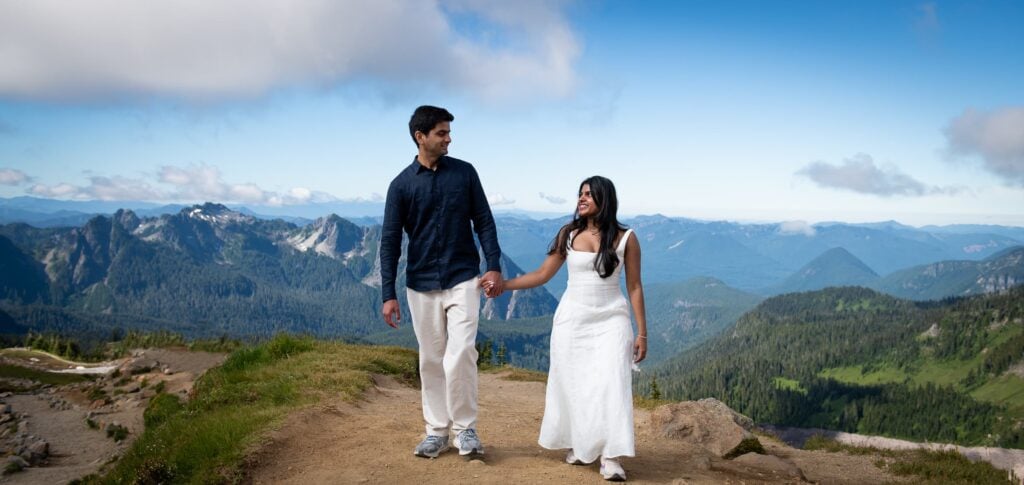 Couple holding hands and walking up trail while looking at each other. Tatoosh range in the background